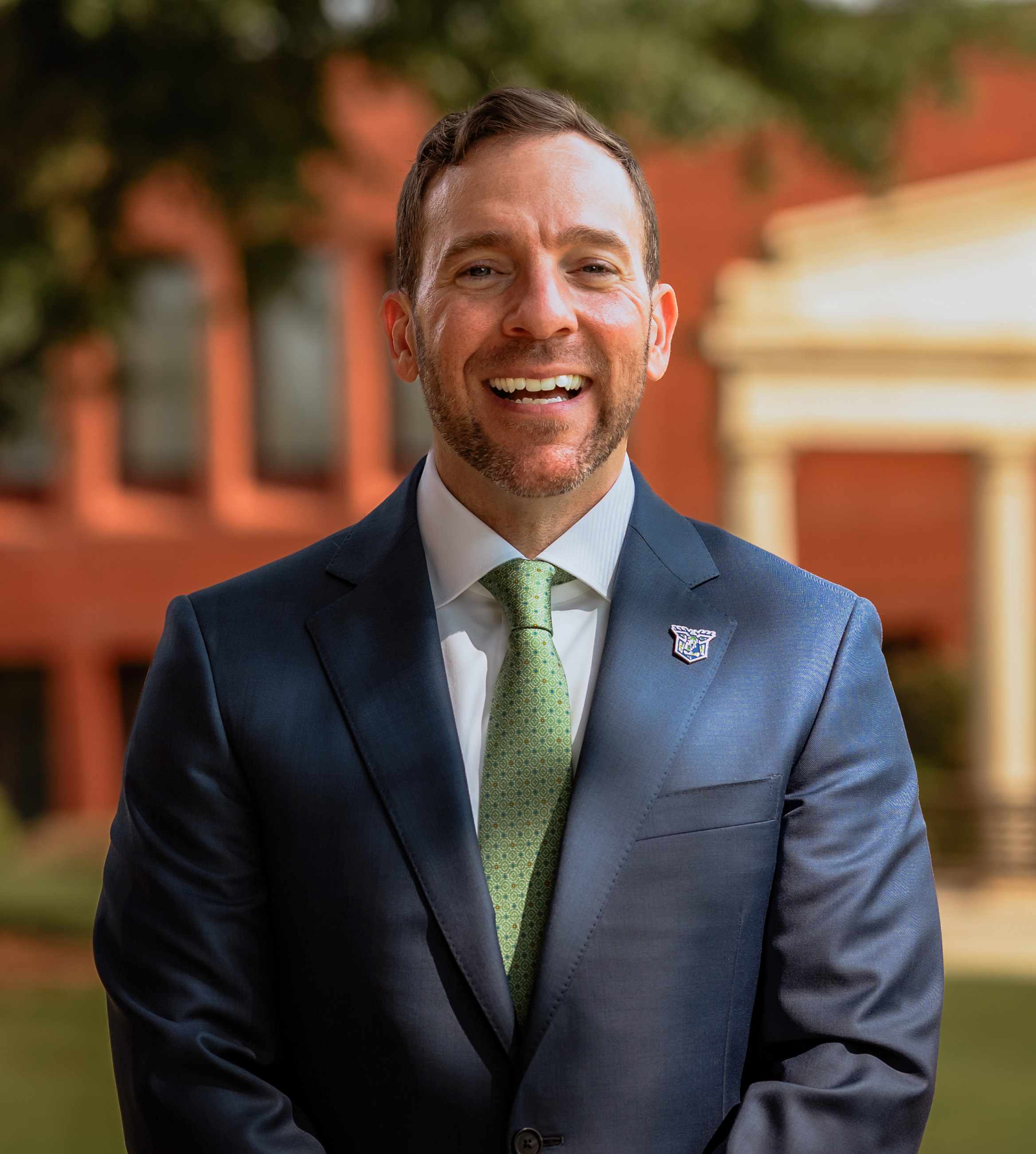 A man in a dark suit and green tie stands outdoors, smiling, with a brick building and greenery in the background.